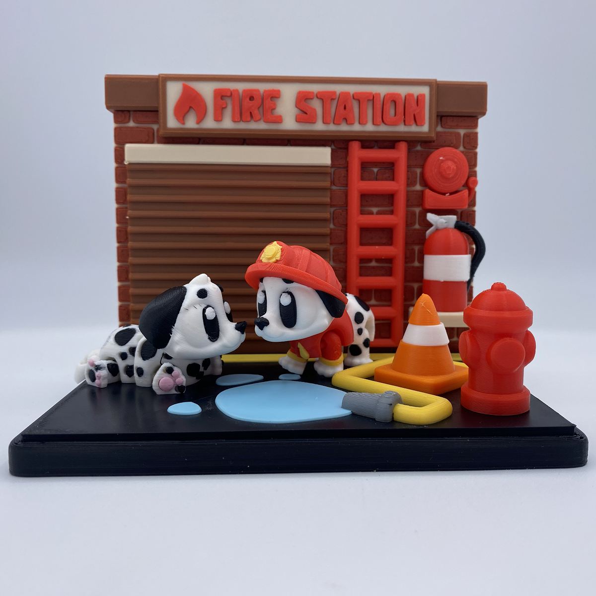A small fire station themed miniature scene with two Dalmatian figures, one wearing a red firefighter helmet and the other lying beside a blue puddle, hose, traffic cone, red fire hydrant, and a brick-style building labeled FIRE STATION.