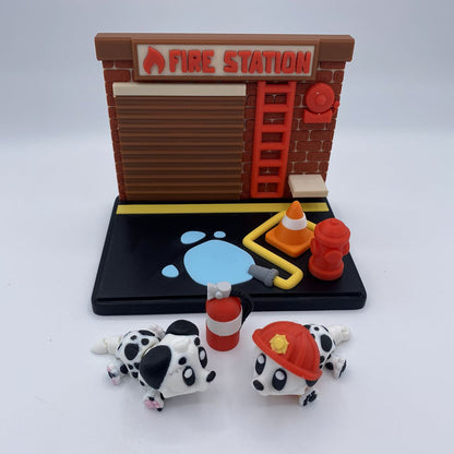 A toy fire station playset with a brown brick-style station building, black base, red fire hydrant, yellow hose, orange traffic cone, blue water splash, and two small Dalmatian figurines in front.
