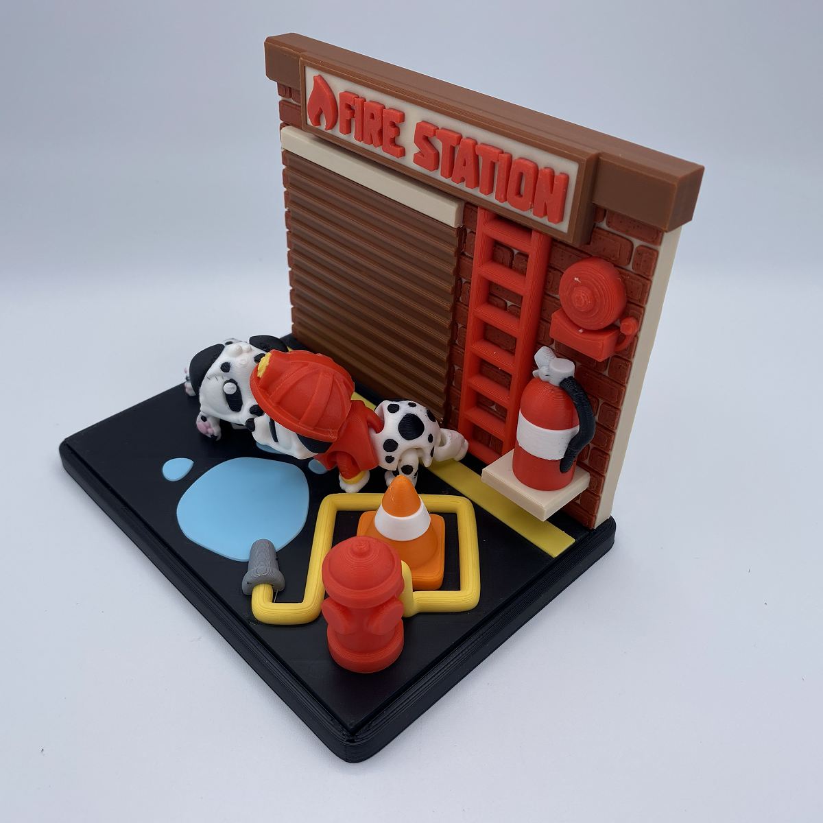A miniature fire station diorama set on a black base, featuring a brown garage door, red ladder, red fire hydrants, a yellow water supply, a blue puddle, and two small dalmatian figurines wearing red firefighter helmets.
