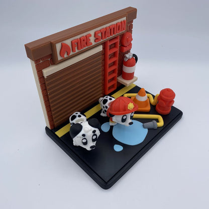 A miniature fire station scene on a black base, featuring a brown brick-style station with a red 'FIRE STATION' sign, a red ladder, two Dalmatian figurines, a firefighter helmet, hydrants, a cone, and a blue puddle detail.