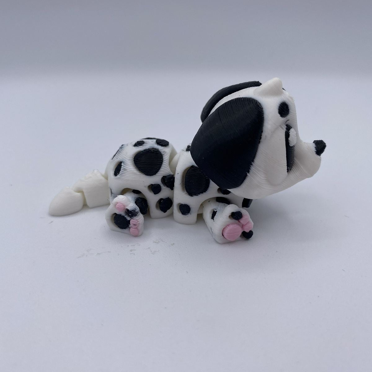 A small white and black spotted dog figurine with pink paw pads, posed on a plain light gray background.