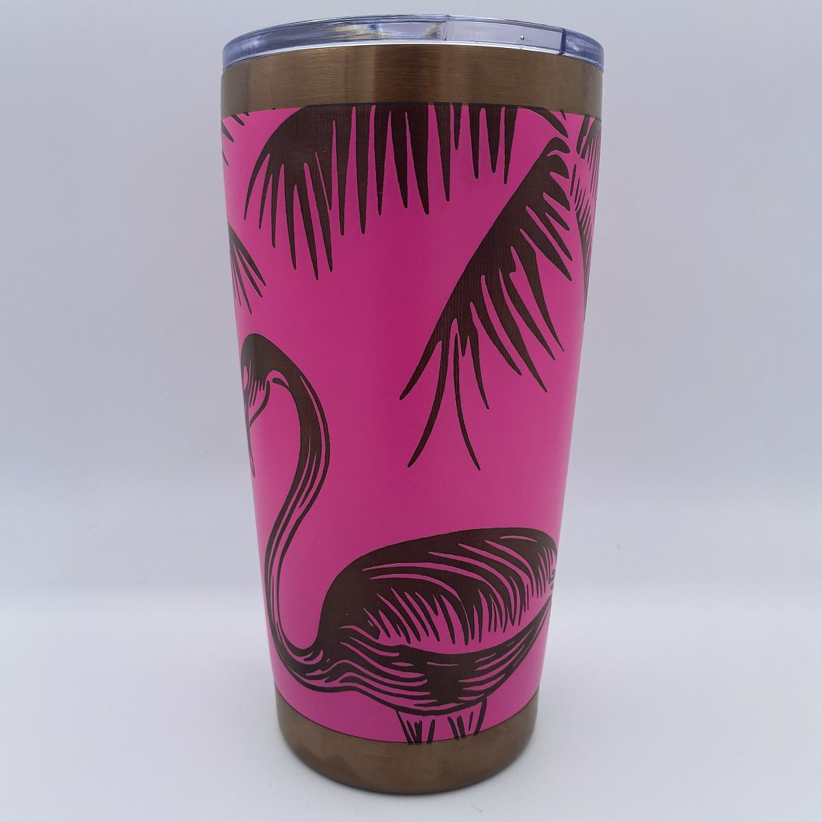 A stainless steel tumbler with a bright pink wrap featuring a flamingo and palm tree illustrations, topped with a clear lid and metallic bronze rim and base.