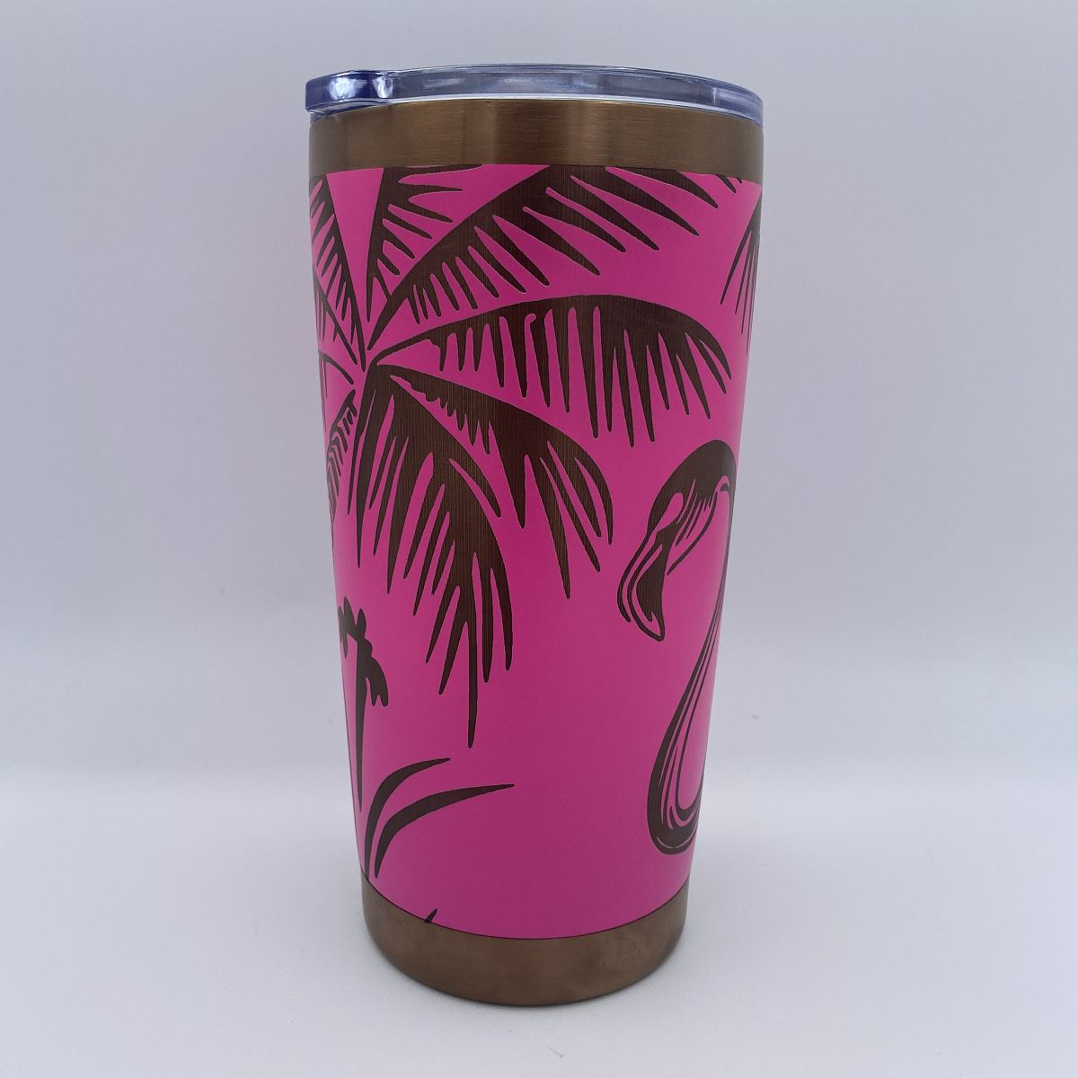 A stainless steel travel tumbler with a bright pink wrap featuring a palm tree and flamingo designs, with a clear lid and metallic bronze rim and base.