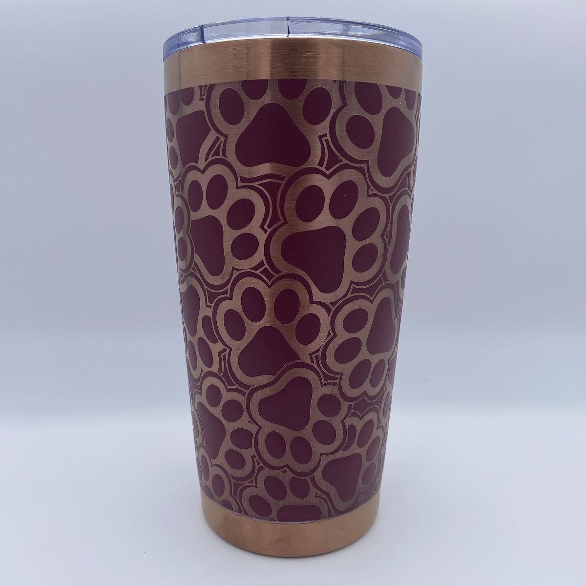 A stainless steel tumbler with a copper-colored rim and base, featuring an all-over paw print pattern in dark burgundy and rose gold. It has a clear plastic lid and is photographed against a neutral light gray background.