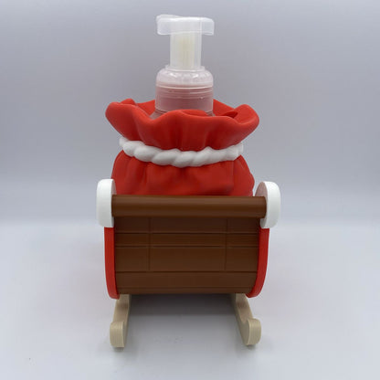 A novelty Christmas-themed foam bottle shaped like a Santa sack in a wooden sleigh, photographed against a neutral gray studio background.
