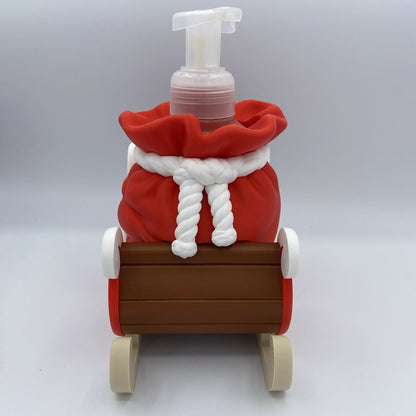 A novelty soap dispenser shaped like a red Santa sack tied with a white rope, sitting in a brown sleigh with beige wheels, on a light gray background.