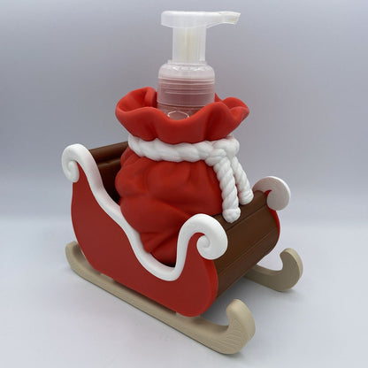 A novelty soap dispenser shaped like a Santa sack in a sleigh, featuring a red body, white trim, beige tracks, and a clear pump bottle on a light gray background.