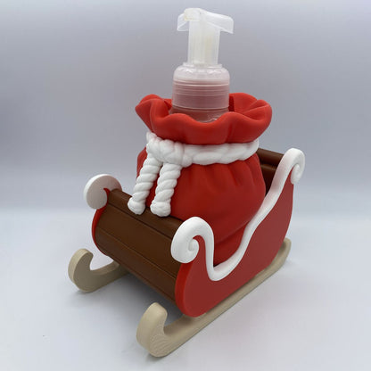 A Christmas-themed foam hand sanitizer dispenser shaped like Santa's sleigh, featuring a red sack with white trim and rope details, a brown wooden-textured body, and beige sleigh wheels, with a clear pump bottle inserted at the top.