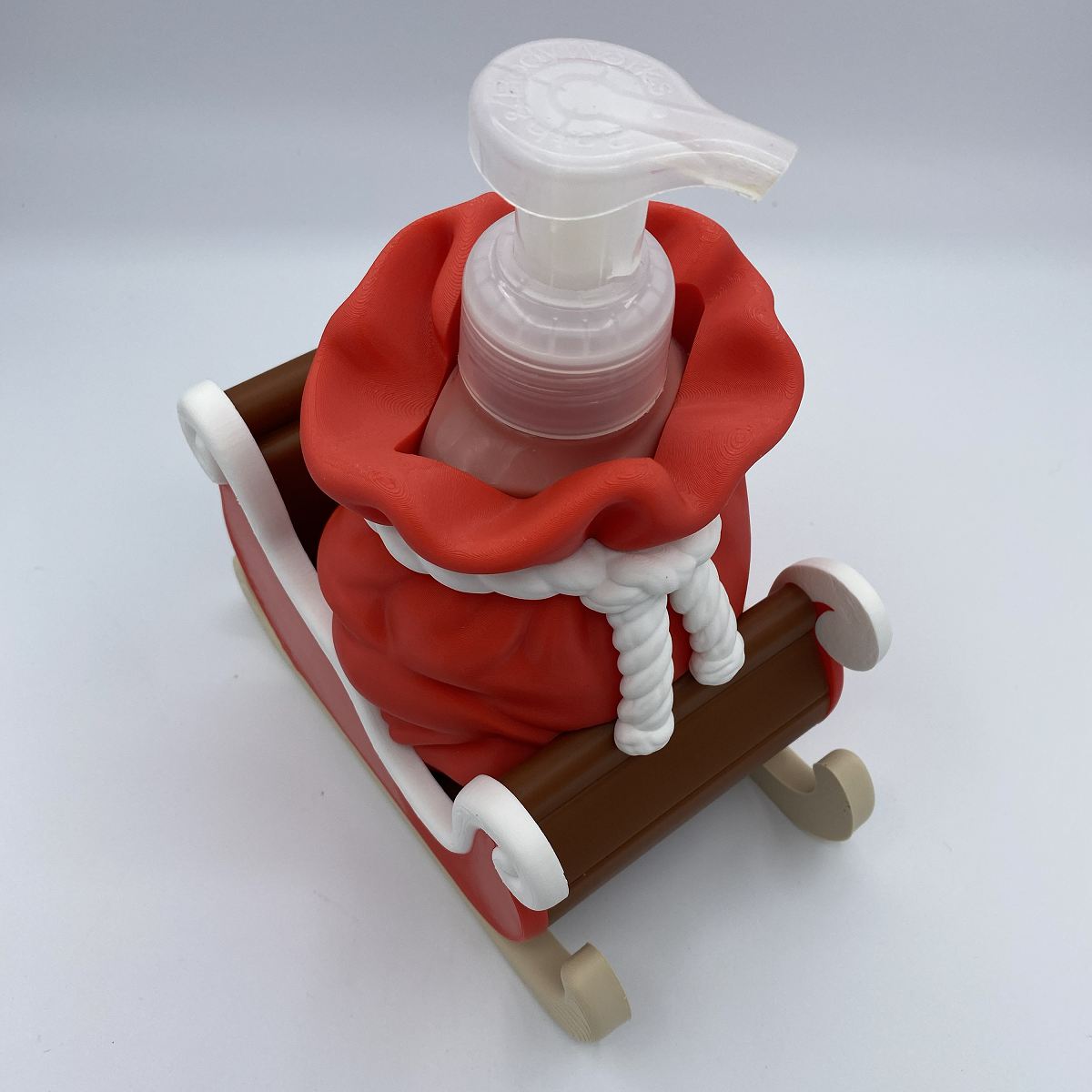 A small Santa-themed soap dispenser shaped like a red sack of soap in a brown sleigh, with a white pump top, photographed on a clean light gray studio background.