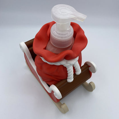 A small Santa-themed soap dispenser shaped like a red sack of soap in a brown sleigh, with a white pump top, photographed on a clean light gray studio background.
