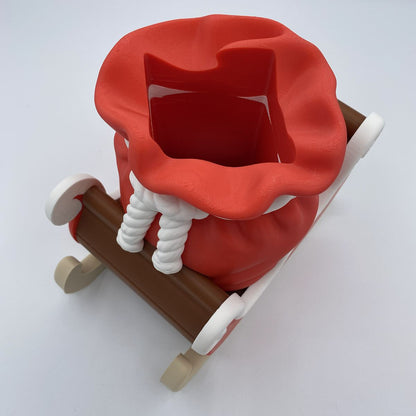 A red Santa sack toy with white rope details and a brown sleigh base, photographed on a clean white background.